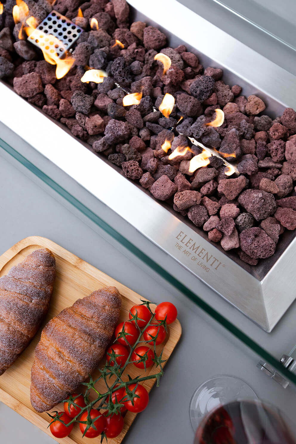 A front angled view of the Elementi Granville Fire Table showing its smooth light grey glass fibre reinforced concrete body with a crisp rectangular silhouette. Centred on the tabletop is a long stainless steel burner pan filled with dark brown and charcoal-coloured lava rock, with bright amber flames rising in an even line. The table rests on a recessed plinth base, creating a subtle floating effect. The background is a clean white studio-style setting which highlights the table’s minimalist modern profile.