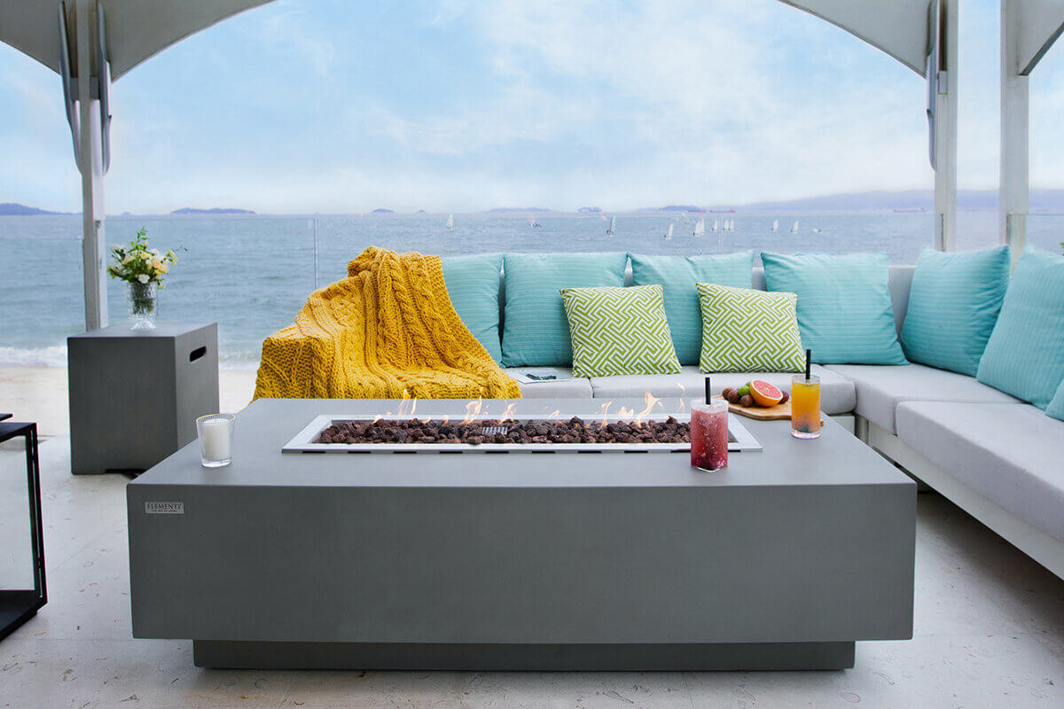 The Elementi Granville Fire Table placed on an outdoor waterfront patio with a sectional sofa dressed in soft turquoise cushions and patterned green accent pillows. A thick mustard yellow knitted throw drapes over the left side of the seating. On the tabletop, bright orange flames appear above the bed of lava rock. A white candle rests at the front left corner while two drinks with black straws sit near the right side. Behind the fire table, the calm ocean stretches toward a hazy horizon with small sailboats scattered across the water.