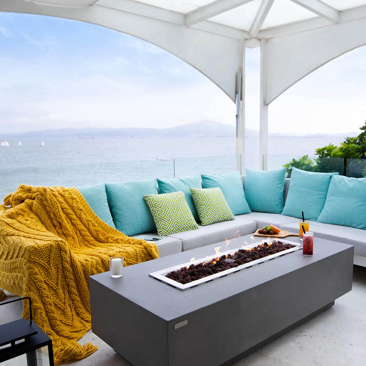 A square-cropped seaside patio scene featuring the Elementi Granville Fire Table centred in front of a curved sectional sofa with turquoise cushions and bold green geometric pillows. The fire is lit, with a strong line of flames rising above reddish black lava rock. A yellow knitted throw blankets the left side of the seating. A pink iced drink and a bright yellow drink sit on the right side of the table on a wooden tray with sliced grapefruit and limes. Soft ocean waves and a pale blue sky form the backdrop.