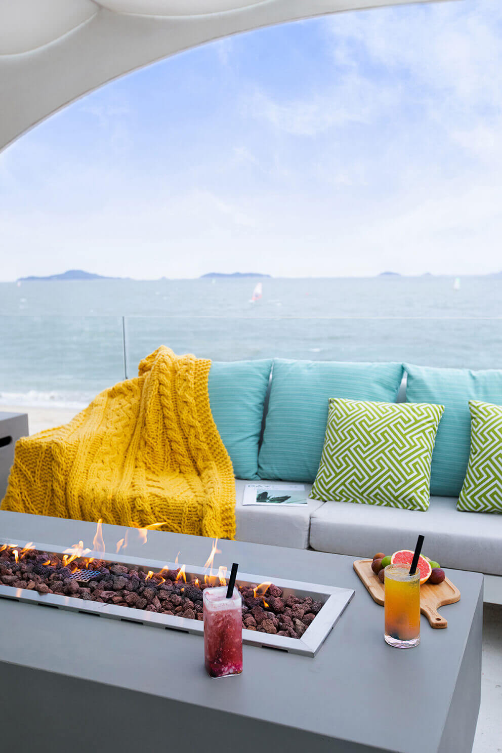 A tall vertical view of the Elementi Granville Fire Table positioned on a seaside terrace. The fire is fully lit, with dancing orange flames above rough lava stones. In the foreground, a red iced drink with a black straw sits on the front right corner of the fire table and a yellow drink rests further back on a wooden tray with citrus fruit. The turquoise and green cushions of the L-shaped sofa create a bright setting, and the ocean beyond the glass railing shows scattered sailboats under a clear pale sky.