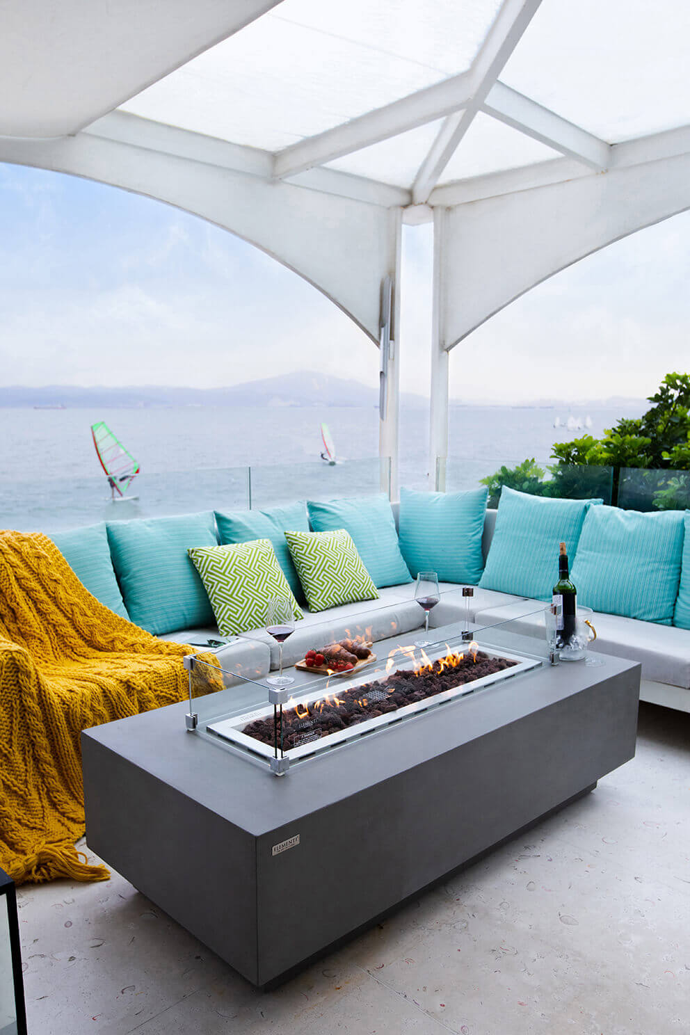 A light grey Elementi Granville Fire Table made from smooth glass fibre reinforced concrete sits on a covered outdoor patio overlooking a bright blue waterfront. The rectangular table features a long central burner tray filled with dark lava rock, protected by a transparent glass wind guard secured by metal corner brackets. Flames rise evenly along the full burner length, casting warm reflections onto the polished glass panels. The table is placed in front of a white outdoor sectional with turquoise and lime green patterned cushions, a yellow knitted throw draped over the left side and two glasses of red wine resting on the table’s right corner. Beyond the seating area, windsurfers glide across the water with hazy mountains in the background, enhancing the relaxing coastal atmosphere. This scene highlights the Granville Fire Table’s modern rectangular silhouette, stainless steel burner components and suitability for propane and natural gas setups.