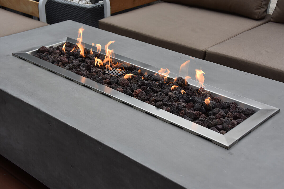 A close angled view of the Elementi Granville Fire Table focuses on its stainless steel burner tray inset into the smooth rectangular grey concrete top. The burner is filled with irregularly shaped dark lava rocks, through which bright flames emerge in a continuous row. The concrete surface shows subtle natural mottling and a fine handcrafted texture. Adjacent outdoor seating with neutral taupe cushions appears softly out of focus in the background, highlighting the table’s modern design and long burner layout. This detail shot showcases the durability of the stainless steel burner components and the included lava rock media.