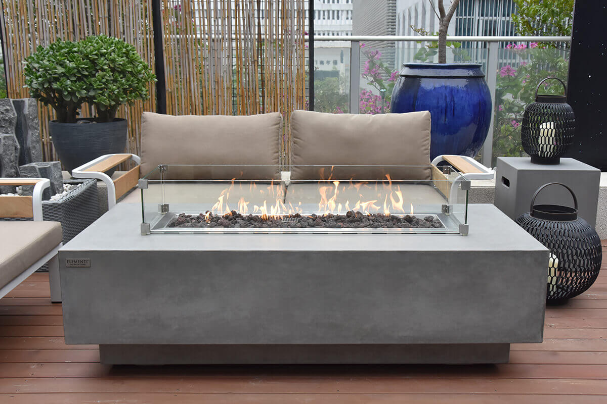 The light grey Elementi Granville Fire Table is positioned on a rooftop-style patio with wooden flooring and modern neutral-coloured seating. The fire table supports a rectangular glass wind guard secured with metal corner brackets, surrounding the long burner filled with dark lava rock. Flames rise cleanly from the centre, casting warm reflections across the glass panels. Surrounding décor includes a tall woven lantern with a pillar candle on the right and a blue ceramic planter with lush greenery behind the seating. The table’s smooth concrete finish and recessed base create a stylish, contemporary centrepiece suitable for propane or natural gas use.