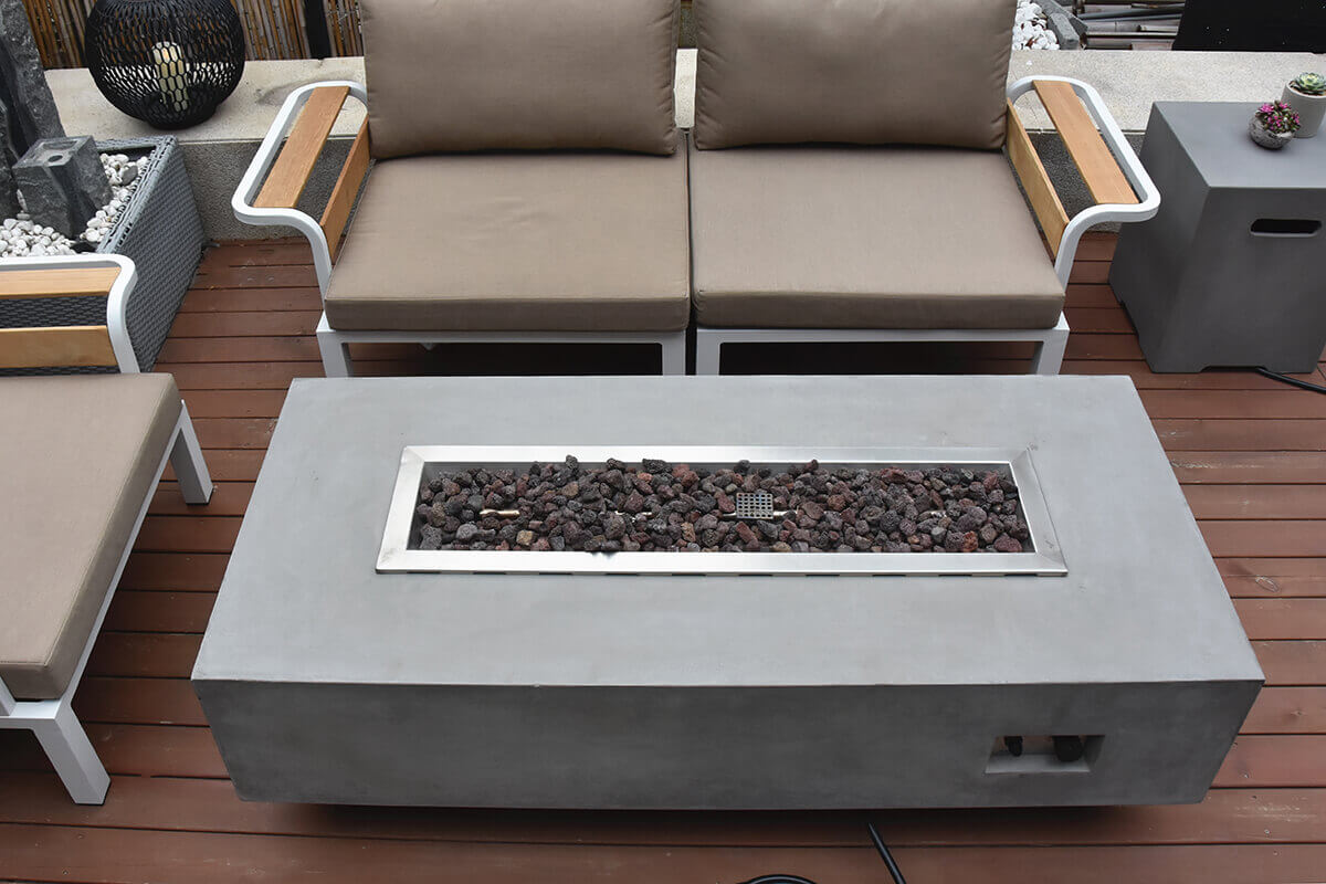 A top-down view of the Elementi Granville Fire Table highlights the full length of its recessed stainless steel burner tray, which is filled with dark lava rock arranged evenly throughout. The rectangular concrete tabletop in light grey shows natural tonal variations and clean straight edges. Outdoor seating with beige cushions and white metal frames surrounds the table, emphasising its proportions and generous burner length. The image provides a clear look at the burner layout, media distribution and handcrafted glass fibre reinforced concrete surface texture.