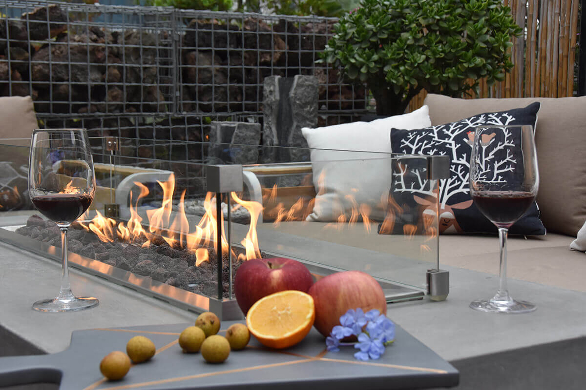 A close-up, intimate view of the Elementi Granville Fire Table shows glasses of red wine positioned in the foreground on the smooth grey concrete surface with the long flame bed extending behind them. In the burner tray, dark lava rocks glow warmly beneath the rising flames, while the surrounding glass wind guard panels reflect the flickering firelight. A cutting board holding apples, an orange half, and small round fruits rests at the front edge, adding a lively social atmosphere. A decorative pillow with a woodland animal design sits on the sofa behind the fire table. This shot highlights the Granville’s ambience, stainless steel burner components and suitability for entertaining.