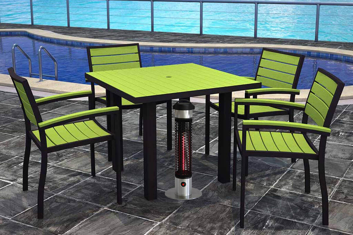 A compact cylindrical HEA21212 Portable Heater standing on a brushed stainless steel round base, positioned in the centre of a square outdoor dining table set beside a bright blue swimming pool. The heater has a black perforated metal grille covering the heating element, which glows with a strong red infrared light running vertically through the core. The top half of the heater is silver with a smooth metallic finish, topped with a black handle-like cap. Four chairs with lime green slatted seats and backs surround the table, evenly spaced on dark stone tiles. The heater sits directly below the table, showing how it provides warmth discreetly from beneath without obstructing legroom.