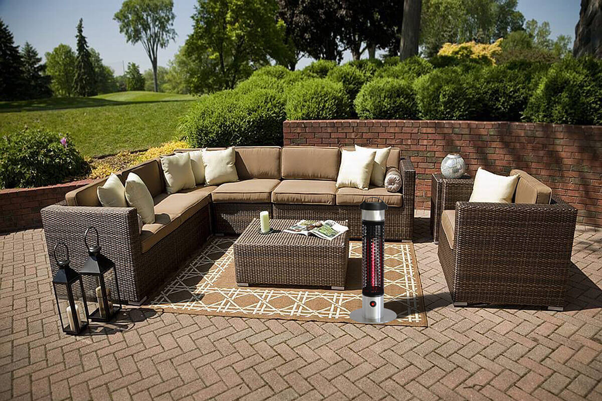 The HEA21212 Portable Heater standing upright on a circular stainless steel base beside a large outdoor wicker sectional sofa set in a landscaped patio area with green lawns and shrubs in the background. The heater’s cylindrical body features a black perforated outer grille revealing a vivid red vertical heating element inside. The upper section is a smooth silver metal cylinder capped with a black top. The patio features brown wicker furniture with beige cushions arranged in an L shape, with a rectangular wicker coffee table in the middle. The compact heater is placed near the table, illustrating its ability to warm seating areas without taking up space.