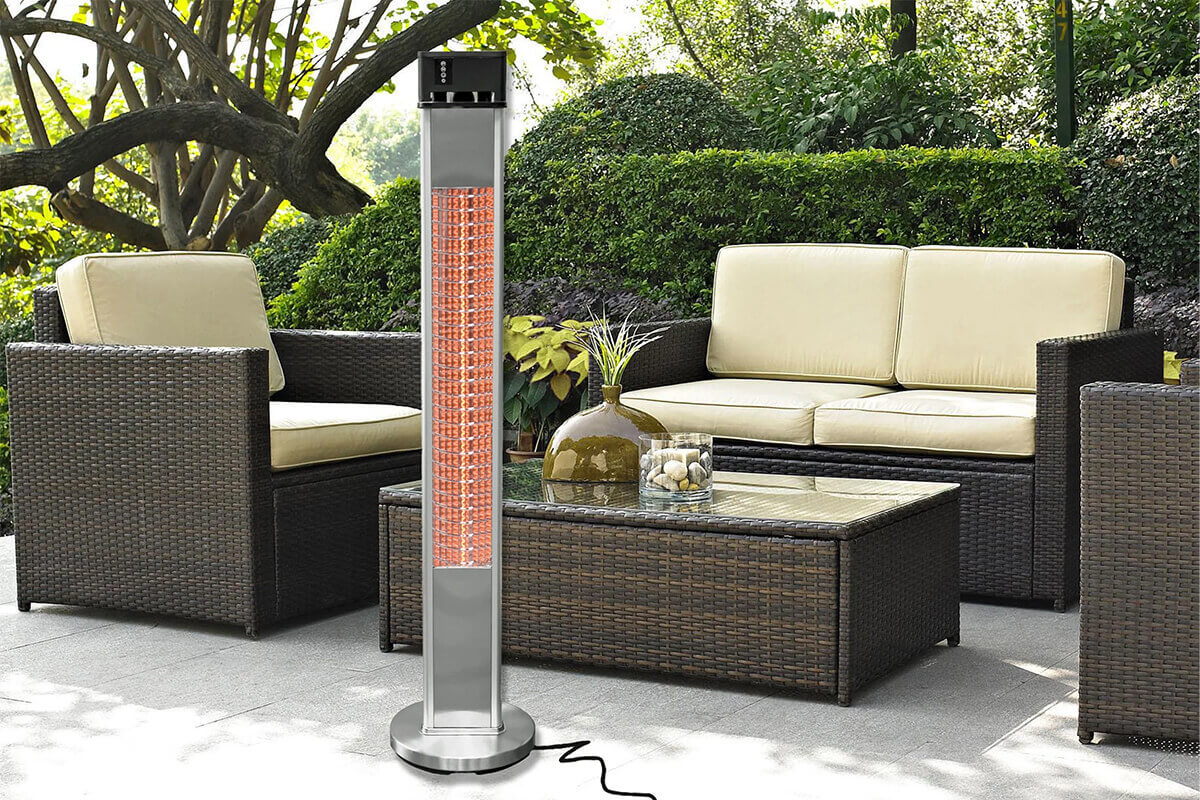 Outdoor patio scene featuring the HEA-215110CVR Standing Infrared Heater positioned on a light grey tiled surface between dark brown woven outdoor furniture. The heater has a slim rectangular brushed-metal column with a glowing orange infrared heating element spanning most of the front face, protected behind a tightly patterned grid. A circular stainless steel base anchors the heater to the ground. A black control housing sits at the top of the unit, with the power cable trailing across the patio. The surrounding setting includes cushioned wicker chairs, a glass-topped coffee table, lush green shrubbery, and mature trees.