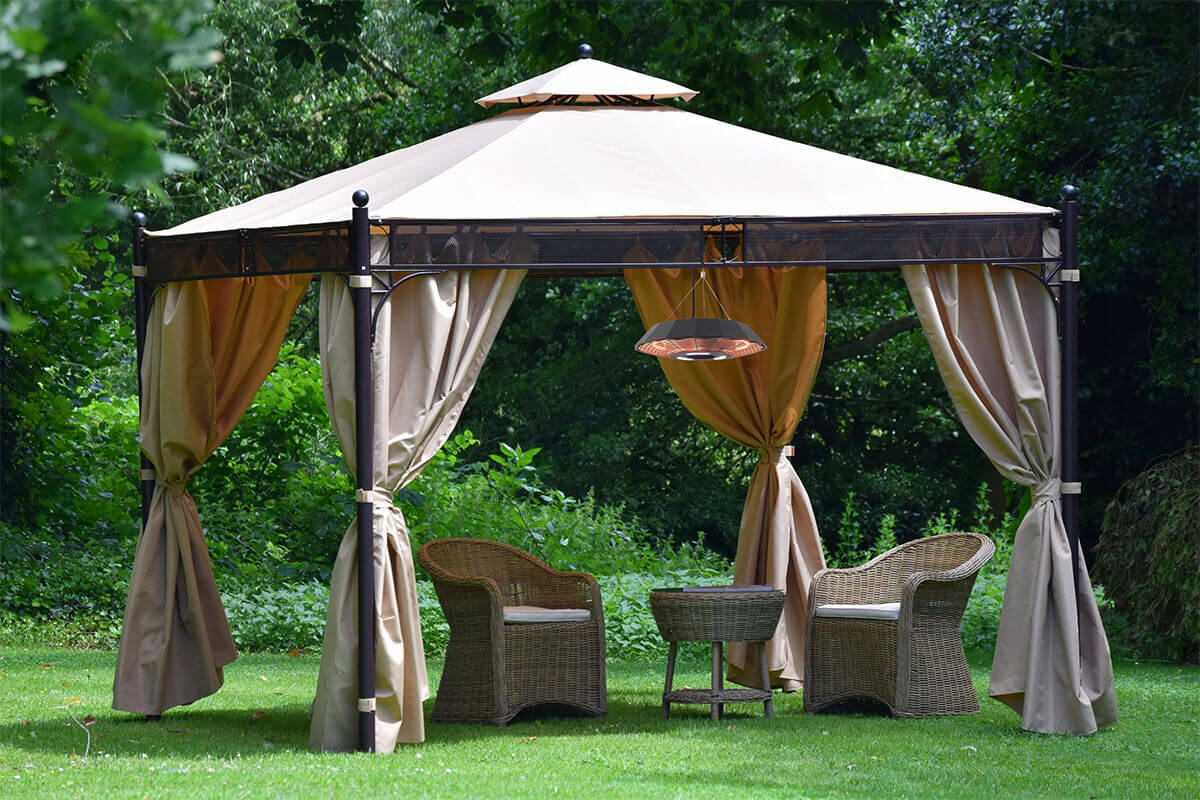Outdoor scene showing the HEA21568 Hanging Infrared Heater suspended from the centre of a large beige gazebo canopy set in a lush green garden. The heater hangs above a small seating arrangement that includes three woven wicker chairs and a matching round wicker table on a neatly cut lawn. The heater’s wide circular housing has a matte dark surface, and its underside features an illuminated ring of orange carbon filament heating tubes protected by a radial metal grille. The warm infrared glow spreads downward onto the table and chairs, while the surrounding trees and dense foliage create a shaded backdrop that contrasts with the heater’s bright warmth.