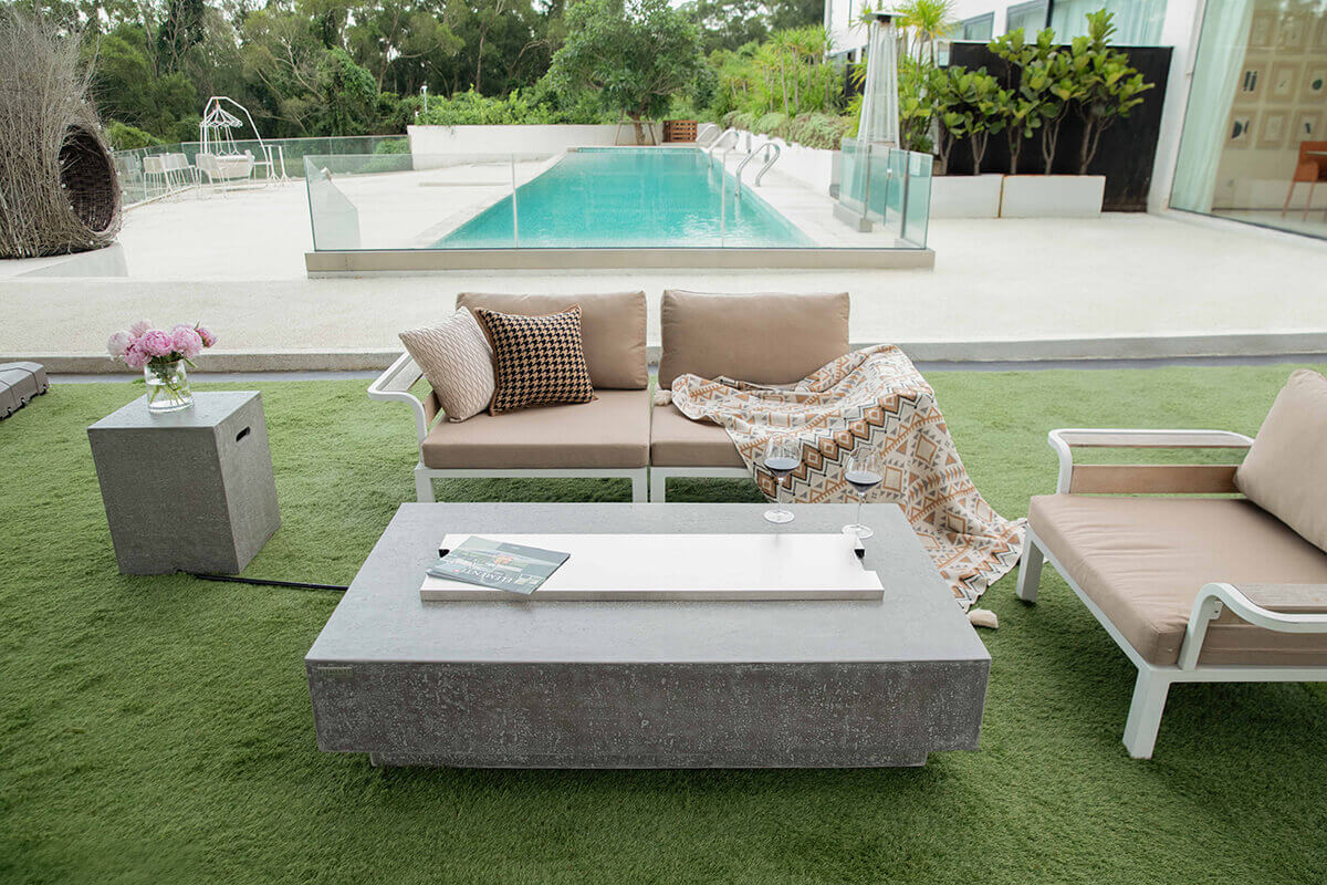 A rectangular Hampton fire table with a light grey glass fibre reinforced concrete finish shown in an outdoor lounge beside a long modern swimming pool. The table’s smooth textured surface holds a removable white burner lid that covers the recessed burner channel. Two stemmed wine glasses rest near the centre edge on the right side of the tabletop. Beige cushioned outdoor seating with patterned throw pillows and a geometric blanket sits behind the table on artificial green turf, with glass railings, lush trees, and a modern white building forming the background.