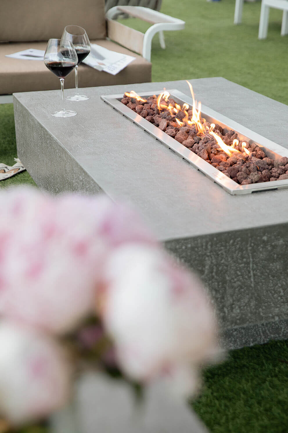 A close angled view of the Hampton fire table showing its stainless steel burner tray filled with rough textured reddish brown lava rocks. Bright yellow and orange flames rise in controlled linear rows along the length of the burner. Two glasses of red wine sit on the tabletop near the upper left corner. The grey concrete surface has a natural pitted texture. Blurred pink flowers appear in the foreground while modern beige seating and green turf form the soft background.