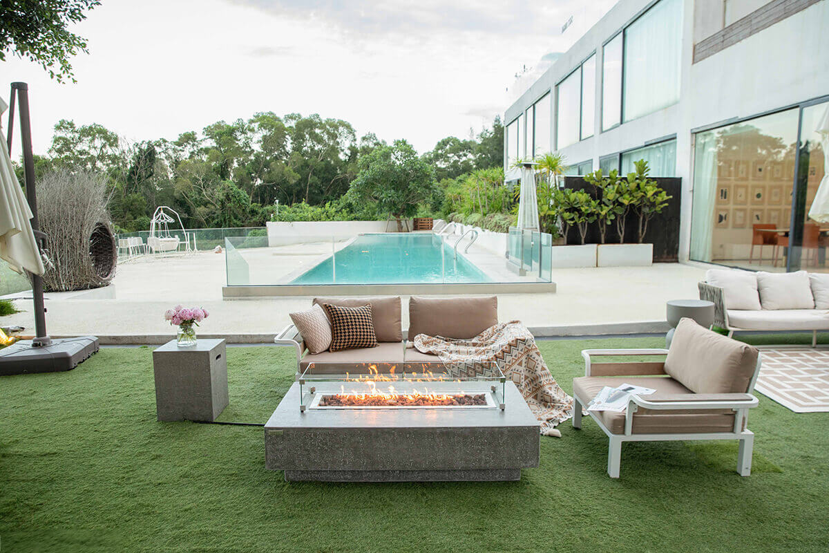 The Hampton fire table positioned in a contemporary outdoor lounge setting beside a long turquoise swimming pool. Flames rise behind the clear glass wind guard, creating a warm glow across the reddish brown lava rocks. The light grey concrete table sits centred between beige cushioned seating with patterned pillows and a geometric throw blanket. A low modern grey side table with a vase of pink flowers stands to the left. Glass railing panels, tall trees, and a minimalist white building complete the scene.