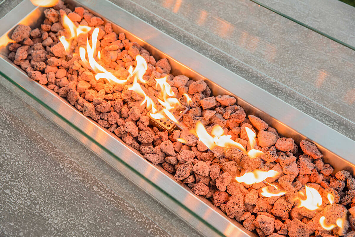 A close close up of the Hampton fire table burner showing bright flames weaving through porous reddish brown lava rocks. The stainless steel burner tray is fully visible along its length, bordered by a clear rectangular glass wind guard. The textured grey concrete tabletop appears at the bottom of the frame, with warm orange flame reflections shimmering across its surface.