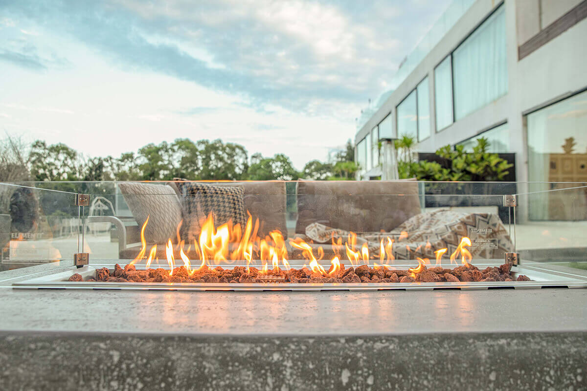 A low front angled view of the Hampton fire table with flames burning behind the glass wind guard. The shot captures the fire at eye level, with the bright orange and yellow flames appearing close against the textured grey concrete tabletop. Behind the table, soft focus beige cushions and patterned throws on the outdoor seating create a warm inviting setting. The tall glass windows of a modern building and lush treetops form the background.