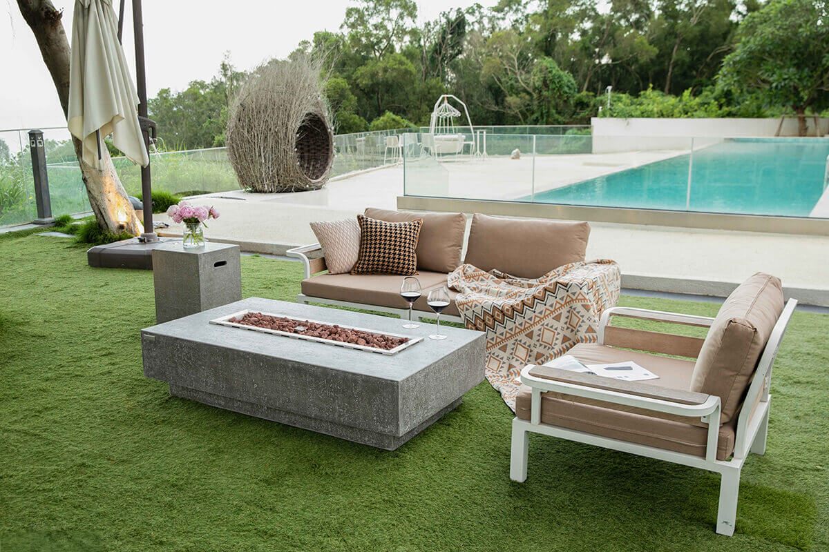 The Hampton fire table placed on green outdoor turf with its stainless steel burner filled with lava rocks. Soft orange flames rise from the centre, protected by a clear glass wind guard. To the left sits a grey side table with a vase of fresh pink flowers. Beige cushioned outdoor seating surrounds the area, and a bright turquoise pool extends into the background behind a glass railing. The scene is calm and resort like, emphasising luxury outdoor living.