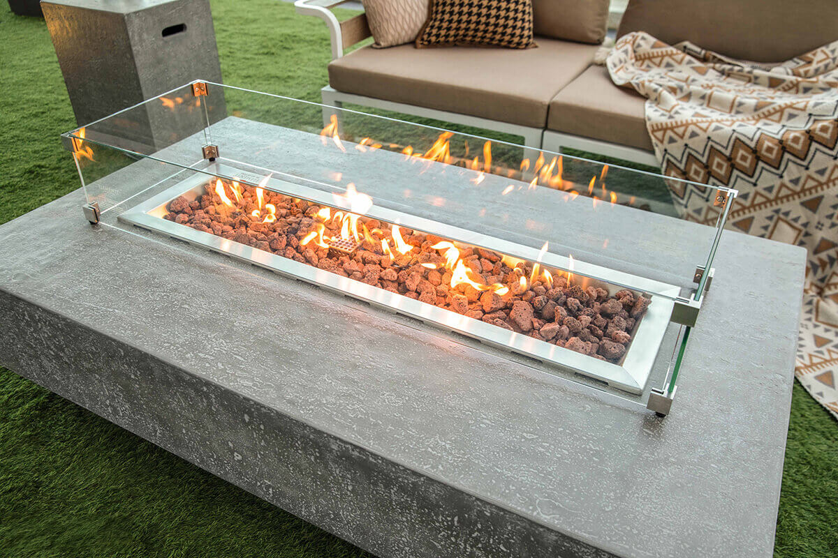 A detailed angled view of the Hampton fire table showing vibrant flames dancing behind the glass wind guard. The stainless steel burner tray holds reddish brown lava rocks arranged in a linear channel. The textured grey concrete tabletop is visible along the bottom edge. Behind the table are beige cushioned seats with patterned pillows and a large geometric blanket draped across them. The soft natural light highlights the clean lines of the modern outdoor lounge.