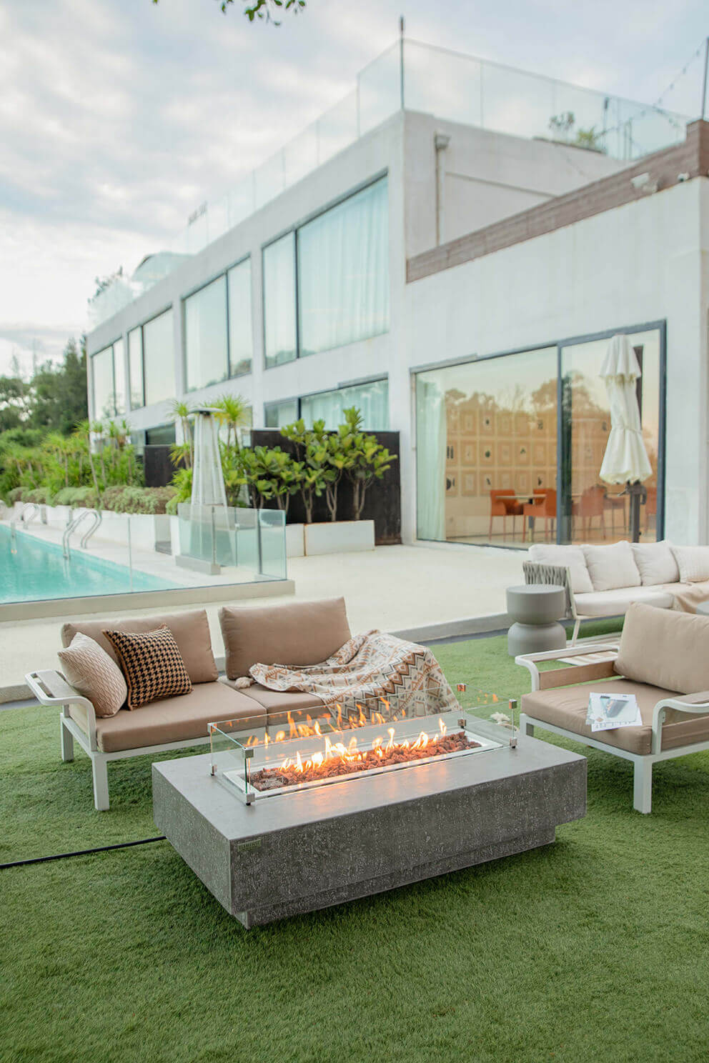 A wide angled view of the Hampton fire table with flames burning brightly across the full length of the stainless steel burner. The table sits on green turf in a modern outdoor living space outside a contemporary white building with large glass windows. Beige cushioned seating surrounds the table. A tall umbrella, potted plants, and the turquoise swimming pool extend toward the left side of the scene, creating a luxurious contemporary atmosphere.