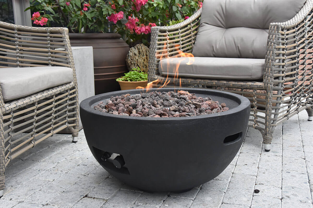 Close-up scene of the Jefferson Fire Bowl in a garden seating area, showing the round black fire bowl filled with textured lava rocks and a small flame rising from the centre, positioned on grey stone pavers between two woven outdoor armchairs with grey cushions, with flowering plants and large planters in the background. The image highlights the fire bowl’s smooth curved rim, recessed control opening, and its suitability for both LP and NG setups.