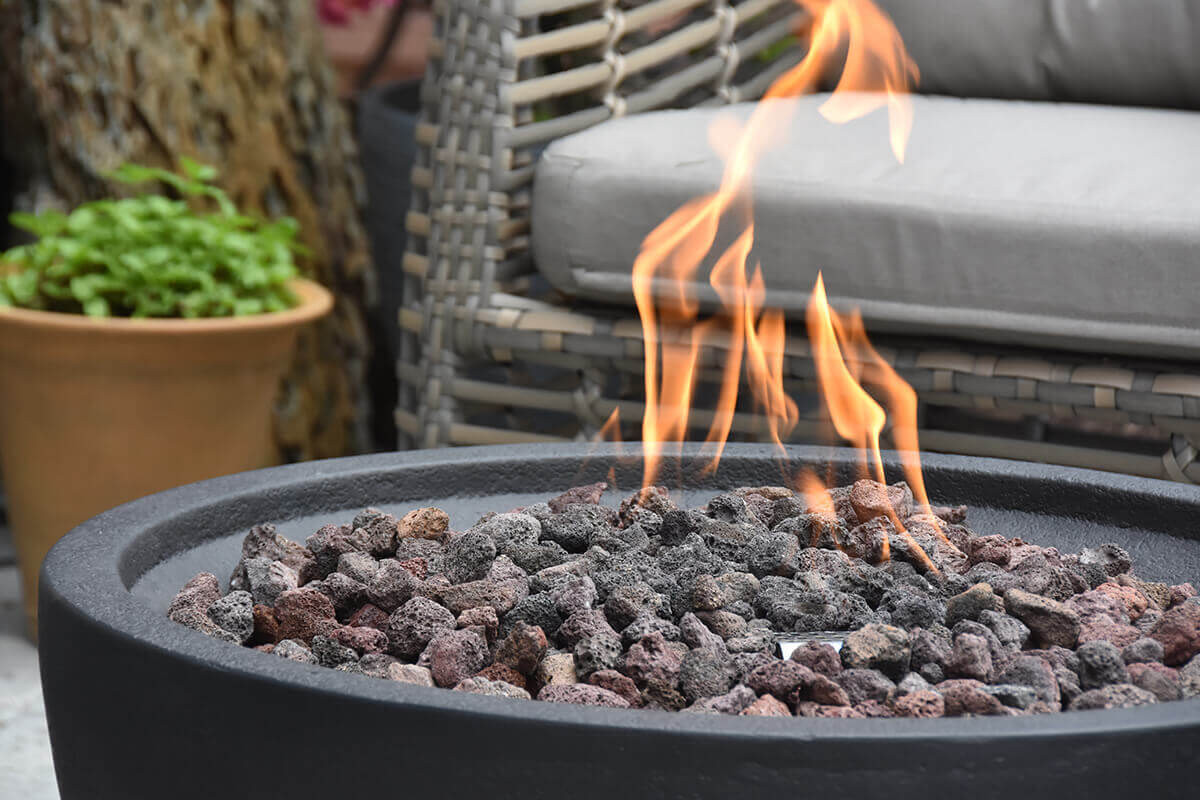 Detailed close-up of the Jefferson Fire Bowl’s burning centre, showing bright yellow-orange flames rising through natural lava rocks of mixed sizes and colours, with part of a woven outdoor chair and grey cushion softly blurred behind. The shot focuses on the fire bowl’s inner basin texture and the heat-resistant burner assembly used for both LP and NG connections.