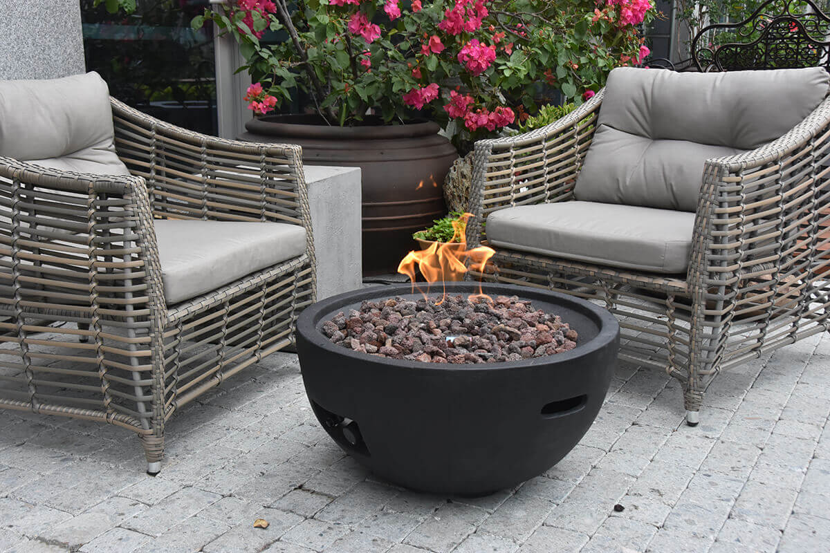 Wide view of the Jefferson Fire Bowl placed on grey stone pavers between two woven outdoor armchairs with grey cushions, surrounded by vibrant pink flowers and large dark planters. The flame burns steadily above the mound of lava rocks, and the bowl’s curved shape, recessed carry handles, and control access slot are clearly visible.