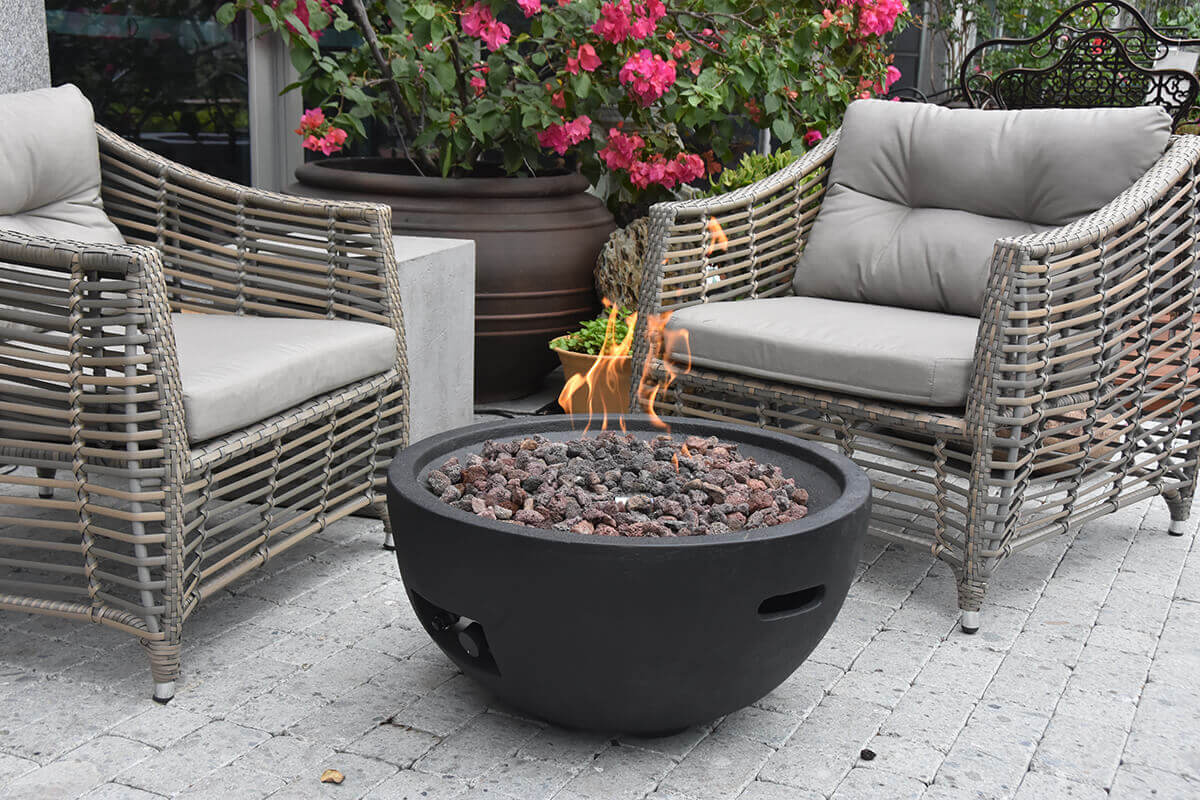 Angled garden-patio view of the Jefferson Fire Bowl with its flame actively burning above a full bed of lava rocks. The scene includes two woven outdoor armchairs and blooming pink flowers, and shows the bowl’s circular form, recessed side handles, textured exterior surface, and functional control opening near the base.