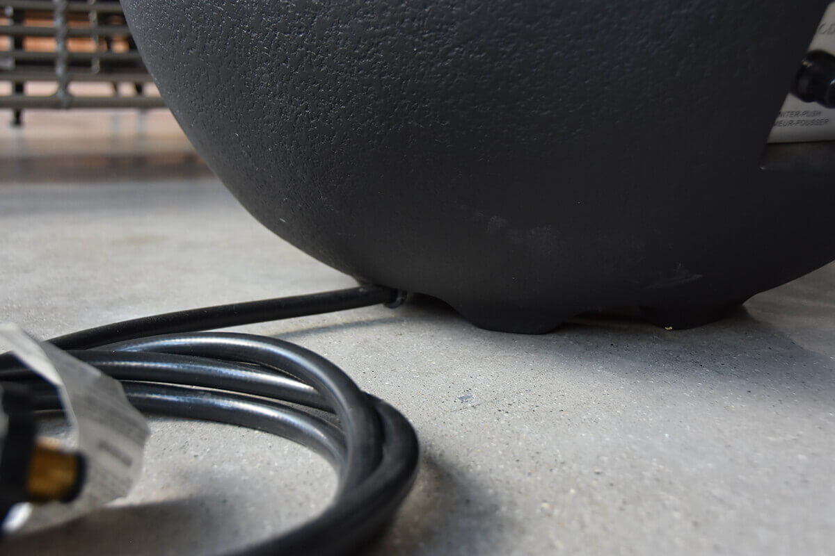 Low-angle close-up of the Jefferson Fire Bowl’s power cord and regulator hose resting on a smooth concrete floor, with the underside curve of the black fire bowl visible above. The image highlights the underside clearance, textured outer finish, and the connection point used for LP or NG fuel systems depending on variation.