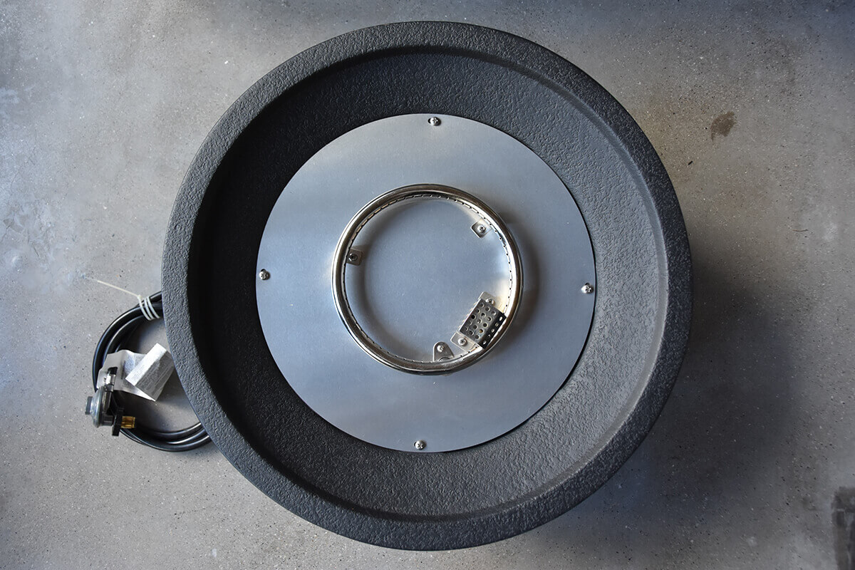 Top-down overhead view of the Jefferson Fire Bowl with all lava rocks removed, revealing the circular burner assembly mounted at the centre of a smooth metal plate inside the bowl. The outer rim shows a textured black finish, and the regulator hose lies to the side. This view clearly displays the burner structure used in both LP and NG fuel configurations.