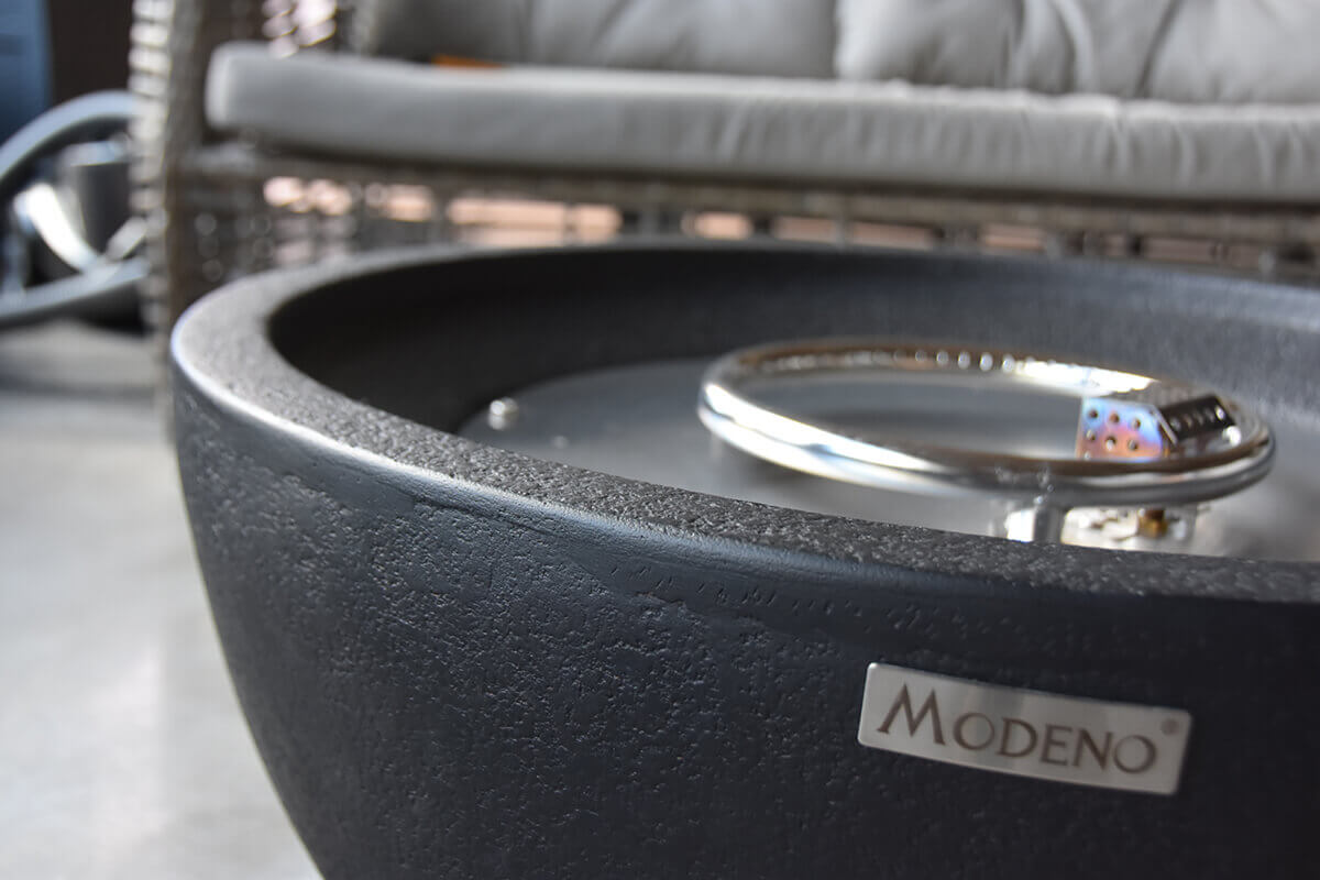 Close-up of the Jefferson Fire Bowl’s upper rim and interior burner area, showing the textured black exterior material curving inward toward the stainless burner ring below. A visible silver emblem labelled “Modeno” is affixed to the outer shell, and soft outdoor seating is blurred in the background.