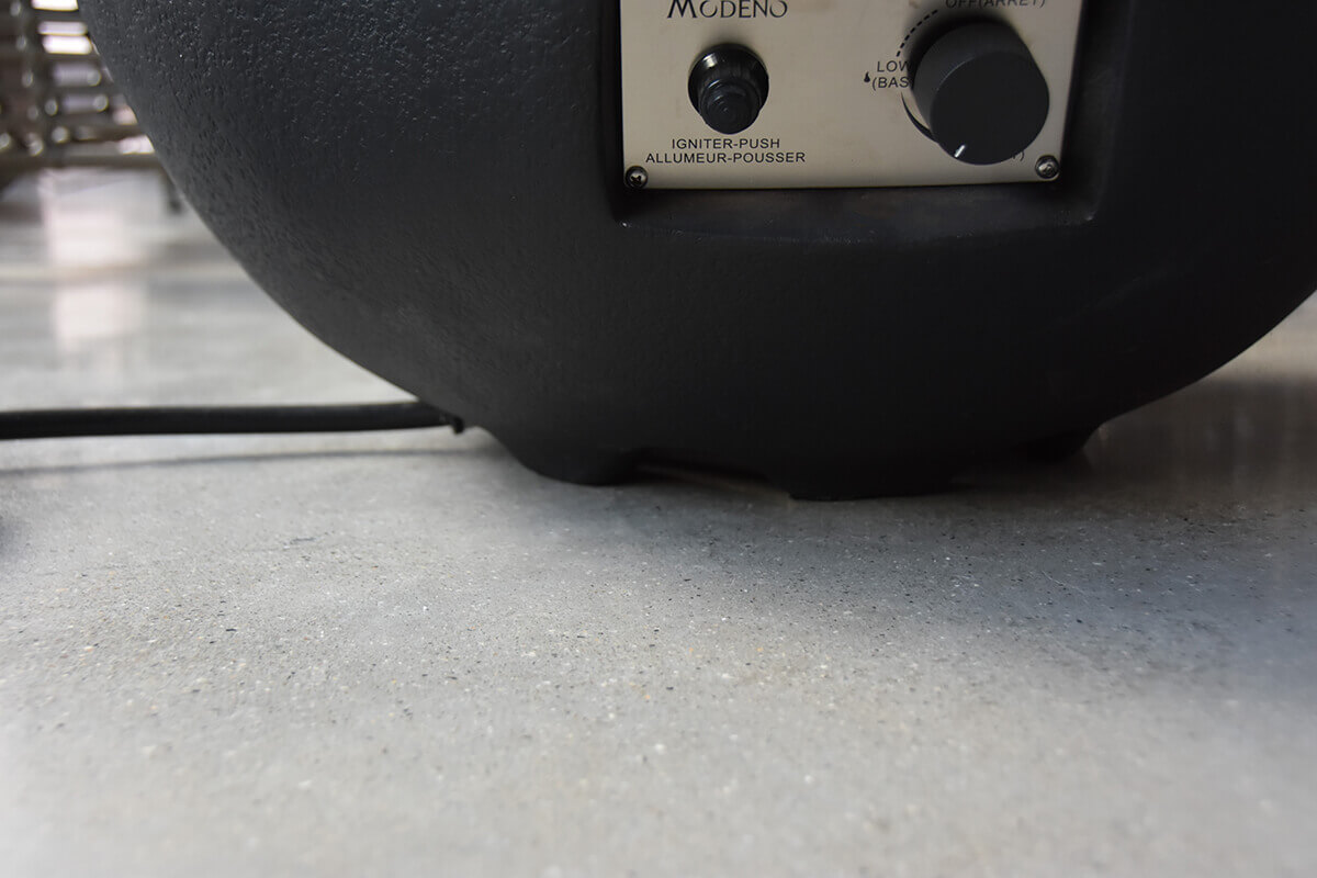 Close-up of the lower side of the Jefferson Fire Bowl, showing the control panel with its push-ignition button and flame adjustment dial inset into a rectangular opening near the base. The textured black surface curves smoothly toward the ground, with part of the regulator hose leading underneath the unit.