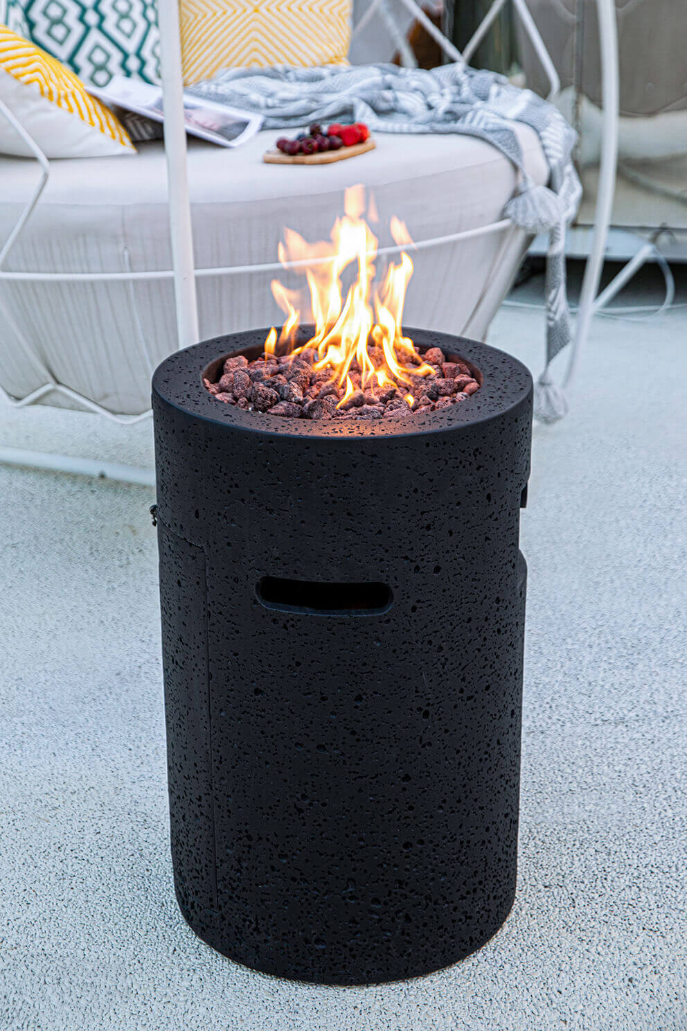 A tall cylindrical black Lava Tube Fire Pit with a heavily textured glass fibre reinforced concrete exterior, shown outdoors beside a cushioned lounger with patterned throw pillows. The burner opening at the top is filled with irregular red and charcoal coloured lava rocks that sit in a recessed circular basin, with tall yellow and orange flames rising from the centre. Two elongated ventilation openings are visible on the upper side of the cylinder, and a control panel cutout sits lower on the left side. Soft evening light highlights the texture of the material while the surrounding patio surface appears smooth and pale.