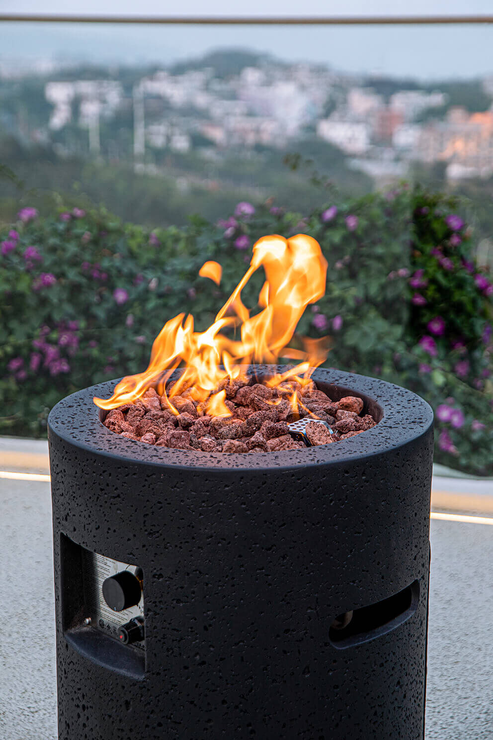 A close view of the Lava Tube Fire Pit on an outdoor terrace with a distant hillside and purple flowering shrubs in the background. The cylindrical black textured exterior rises to a recessed circular burner filled with chunky lava rocks in shades of red and grey. Bright flames emerge from the centre of the rock bed. A long horizontal ventilation opening is visible on the side of the cylinder. The fire feature stands on a light coloured outdoor surface with soft natural light illuminating its textured surface.