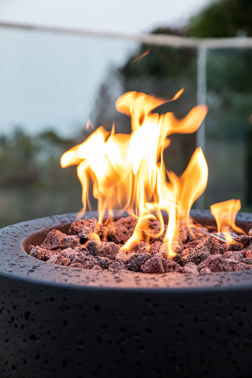 A tight close up of the top of the Lava Tube Fire Pit showing vivid yellow and orange flames rising directly from the uneven lava rock bed. The rocks have a porous texture with a mixture of red, brown, and charcoal tones. The curved inner rim of the burner opening appears dark and textured, consistent with the cylindrical body. The out of focus background reveals glass railing reflections and hints of sky, emphasizing the flame movement and rock detail.