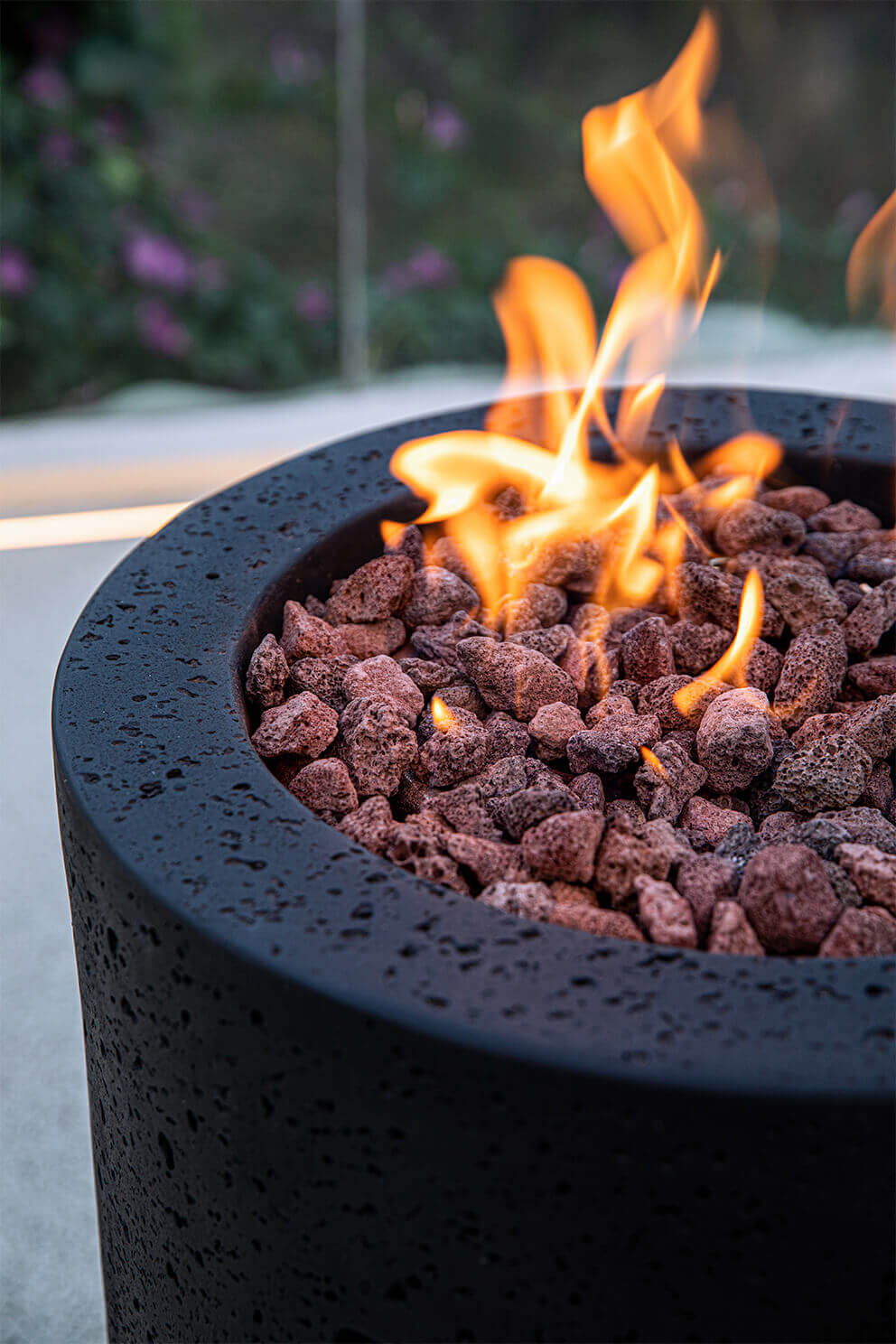 A close angled view of the Lava Tube Fire Pit focusing on the upper section of its textured black cylindrical body. The recessed circular burner is filled with red and brown lava rocks that surround the flame source. Soft orange flames rise from several points within the rock bed. The outer rim of the burner opening is thick and rounded with a speckled texture. The background includes blurred natural greenery and the edge of a patio surface.