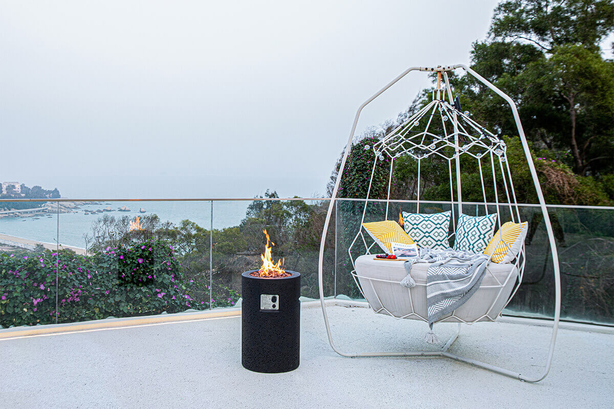 A wide scene of the Lava Tube Fire Pit placed on an elevated patio overlooking a coastline. The black textured cylinder stands beside a suspended white lounge chair with patterned yellow and teal cushions and a grey woven blanket draped across it. The fire feature’s recessed burner glows with visible flame rising from lava rocks. A glass railing runs across the patio edge, framing views of trees, shoreline, and water below under a hazy sky.