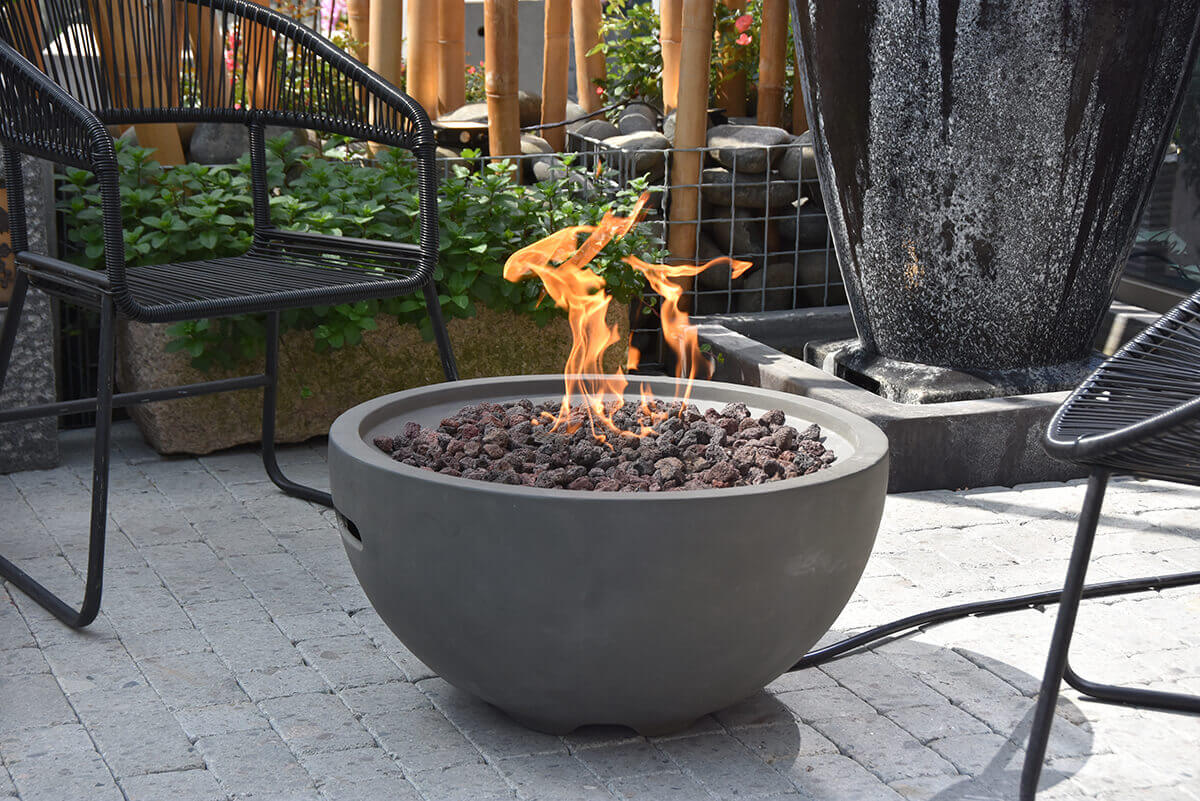 A light grey Nantucket Fire Bowl sits on an outdoor stone patio surrounded by modern black wire chairs and lush green plants. The smooth rounded concrete body has a soft matte finish with subtle surface variations. Bright orange flames rise from a deep bed of chunky red and charcoal coloured lava rocks arranged inside the recessed burner opening. The fire bowl’s curved upper rim is evenly thick and slightly bevelled, framing the central flame area with clean symmetry. In the lower side of the bowl, a narrow horizontal ventilation slot is partially visible. Behind the fire bowl, bamboo posts, stacked stones and a tall dark water feature create a natural backdrop.