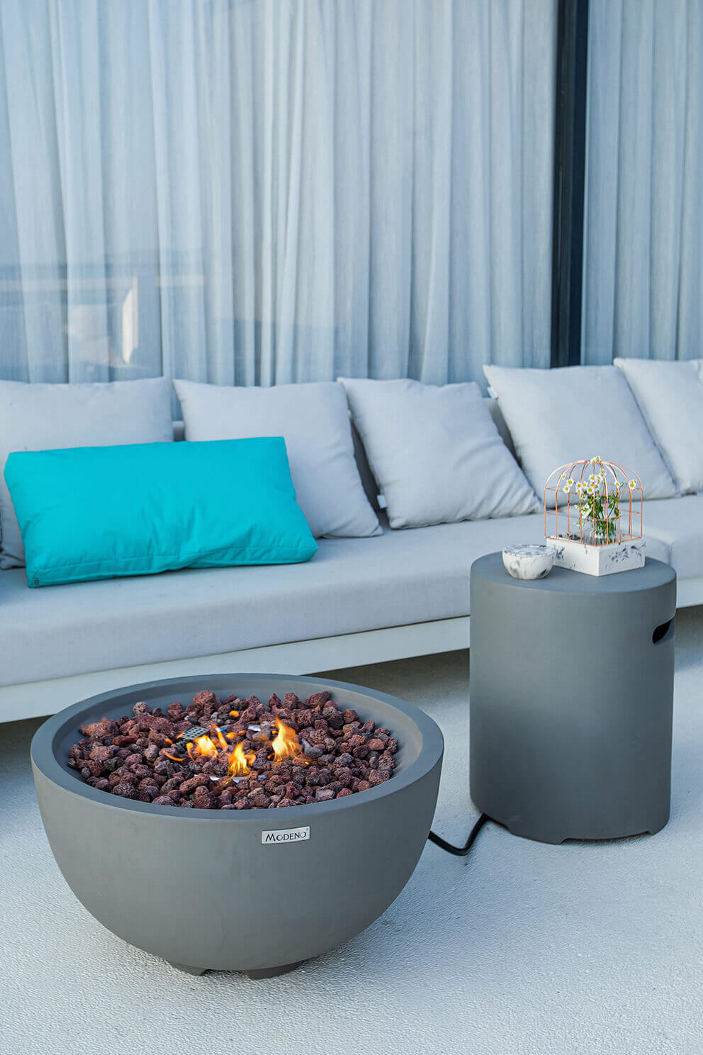 The light grey Nantucket Fire Bowl is positioned in front of a long outdoor sofa with white cushions and a bright teal accent pillow. The bowl’s smooth concrete surface is clearly defined under soft daylight. Gentle orange flames rise from a centre bed of rough lava rocks, which vary in texture and shades of reddish brown and black. A matching grey cylindrical tank cover with a cutout handle stands beside the fire bowl, connected by a visible black hose that trails across the ground. The fire bowl’s aluminium burner ring is subtly visible beneath the lava rock layer, showing an even flame distribution suitable for either LP or NG setups.
