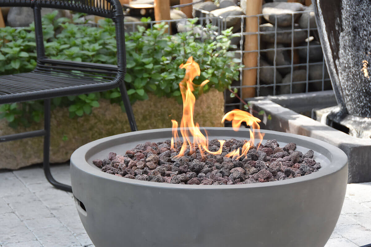 A close view of the flame area of the Nantucket Fire Bowl shows tall curling orange flames emerging from a dense layer of porous lava rocks. The bowl’s light grey concrete rim forms a smooth circular frame around the centre fire pit. The background includes a woven black outdoor chair and dense green foliage, softly blurred, drawing focus to the textured stone-like finish of the fire bowl and the lively movement of the flames. The lava rocks vary in size and colour, from dark charcoal to reddish tones, and sit atop the concealed burner assembly.