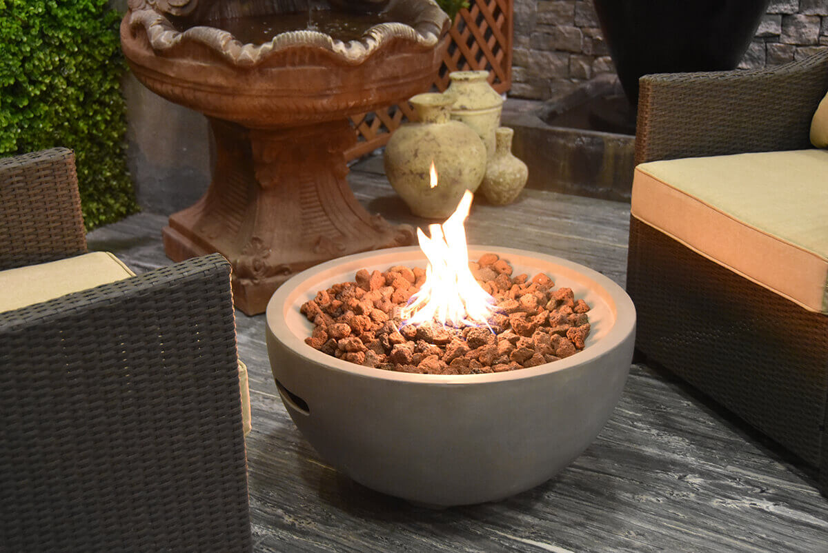 The Nantucket Fire Bowl emits a bright central flame in a warmly lit outdoor seating area. Two wicker armchairs with beige cushions sit on either side of the bowl, framing the scene. The fire bowl’s light grey concrete body has a smooth, gently curved shape that rests on small rounded feet. The bowl is filled with reddish brown and dark grey lava rocks spread evenly across the burner opening. Behind the seating arrangement, a stone wall, large urn, and decorative fountain create an intimate garden atmosphere illuminated by the fire’s glow.