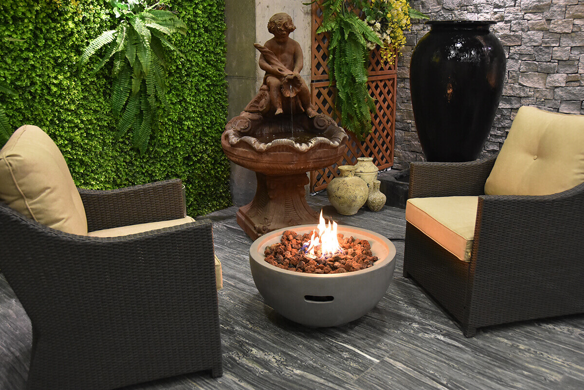 A wider nighttime view shows the Nantucket Fire Bowl centred between two wicker armchairs with yellow-beige cushions. The fire bowl’s flames glow brightly from the circular bed of irregular lava rocks. The light grey concrete body features a single horizontal handle-style opening near the base. The surrounding patio includes a richly textured stone wall, greenery, a decorative water fountain and pottery pieces, creating a cosy enclosed courtyard ambiance. The fire bowl provides a warm focal point suitable for either LP or NG configurations.