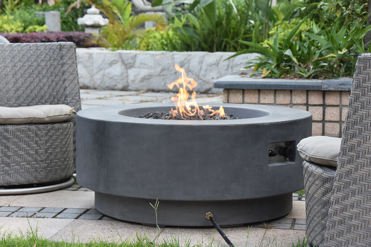 Extremely detailed outdoor scene showing the Ross Fire Table in a landscaped garden, built in a smooth light-grey glass-fibre reinforced concrete with a broad circular top and a recessed fire bowl at the centre. The fire bowl is filled with irregular dark lava rocks surrounding a stainless-steel burner that produces medium-height yellow–orange flames curling upward. The cylindrical body features a clean, modern profile with a slightly recessed lower tier that forms the base. To the right side, a rectangular service opening reveals the control panel with a push-button spark ignition and adjustment knob. Surrounding the fire table are woven grey outdoor chairs with light cushions placed on a stone-paved patio bordered by green grass. The scene highlights both LP and natural gas installation suitability through the visible gas hose connection on the lower edge.
