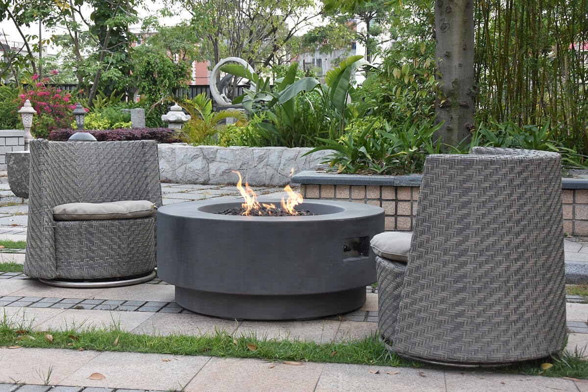 Garden setting with the Ross Fire Table positioned centrally among woven grey patio chairs. The fire table appears in light-grey smooth concrete with a wide circular top and a deep fire chamber containing dark lava rocks. Bright, twisting flames emerge from the stainless-steel burner beneath the rocks. A rectangular access cut-out on the side exposes the burner controls and ignition components. The lower section of the table forms a setback circular base that visually anchors the design. Surrounding the installation are manicured plants, stone retaining walls, decorative garden sculptures, and patterned paving stones, illustrating how the Ross Fire Table functions attractively in both LP and natural-gas configurations.