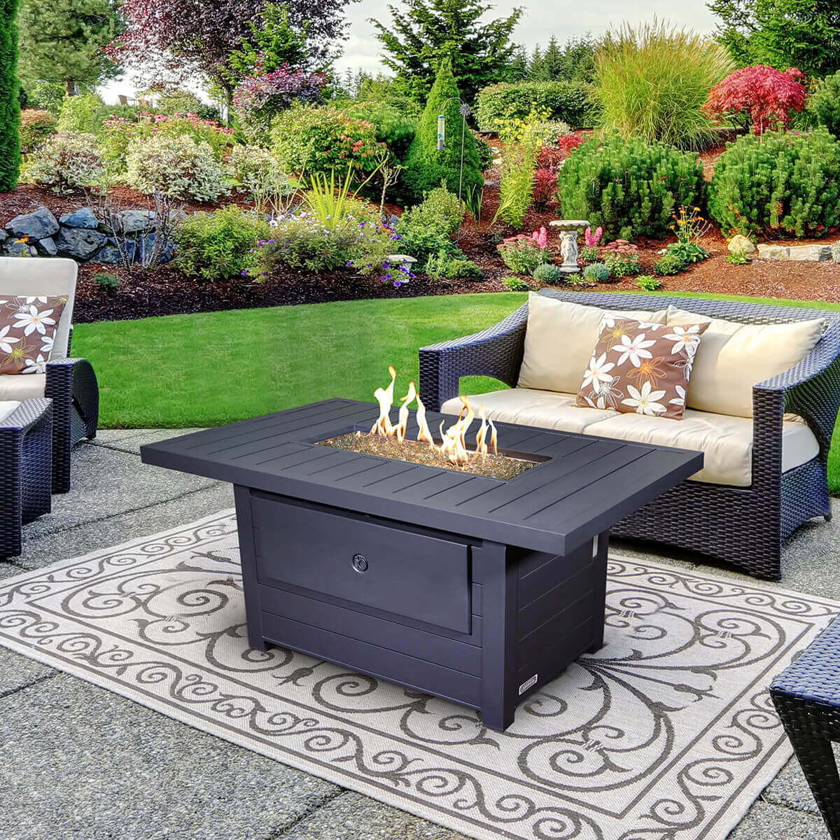 A Sunbeam Serenity Fire Table shown outdoors on a patterned outdoor rug, photographed in soft natural light with the table ignited. The rectangular tabletop has a smooth matte black finish with evenly spaced horizontal slats and a central recessed burner tray filled with clear decorative flame glass that reflects the warm yellow and orange flames flickering upward. The flames rise in uneven peaks across the full length of the burner tray, revealing the stainless steel burner beneath the glass. The surrounding table surface is wide and flat with clean right-angled edges and a seamless join to the sturdy vertical base panels below. The base features a centred rectangular access door that conceals the propane tank, blending into the slatted panel construction. Wicker patio seating with cream cushions surrounds the table, and a well-manicured garden with lush shrubs and flowering plants forms the background, creating a relaxed outdoor setting.