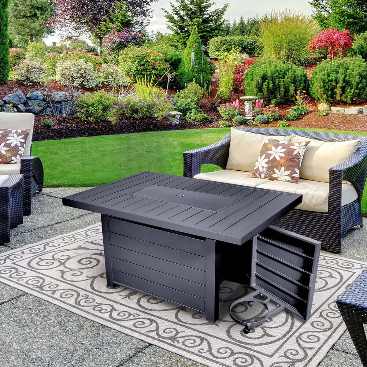 A Sunbeam Serenity Fire Table pictured with the burner area fully covered by the included matching metal lid, converting the table into a continuous flat black surface. The matte tabletop displays its horizontal slat pattern uninterrupted, with the circular lid handle in the centre sitting flush and subtle. The propane tank access door on the front of the base is open, revealing the interior storage compartment and the sliding tank support ring positioned on a low rail system for easy tank loading. The open door shows the neatly finished interior panels and concealed hinges. The table stands on four small feet that provide subtle elevation and stability. The background includes woven patio furniture, a patterned outdoor rug, and a landscaped garden filled with layered shrubs, evergreens, and flowering plants.