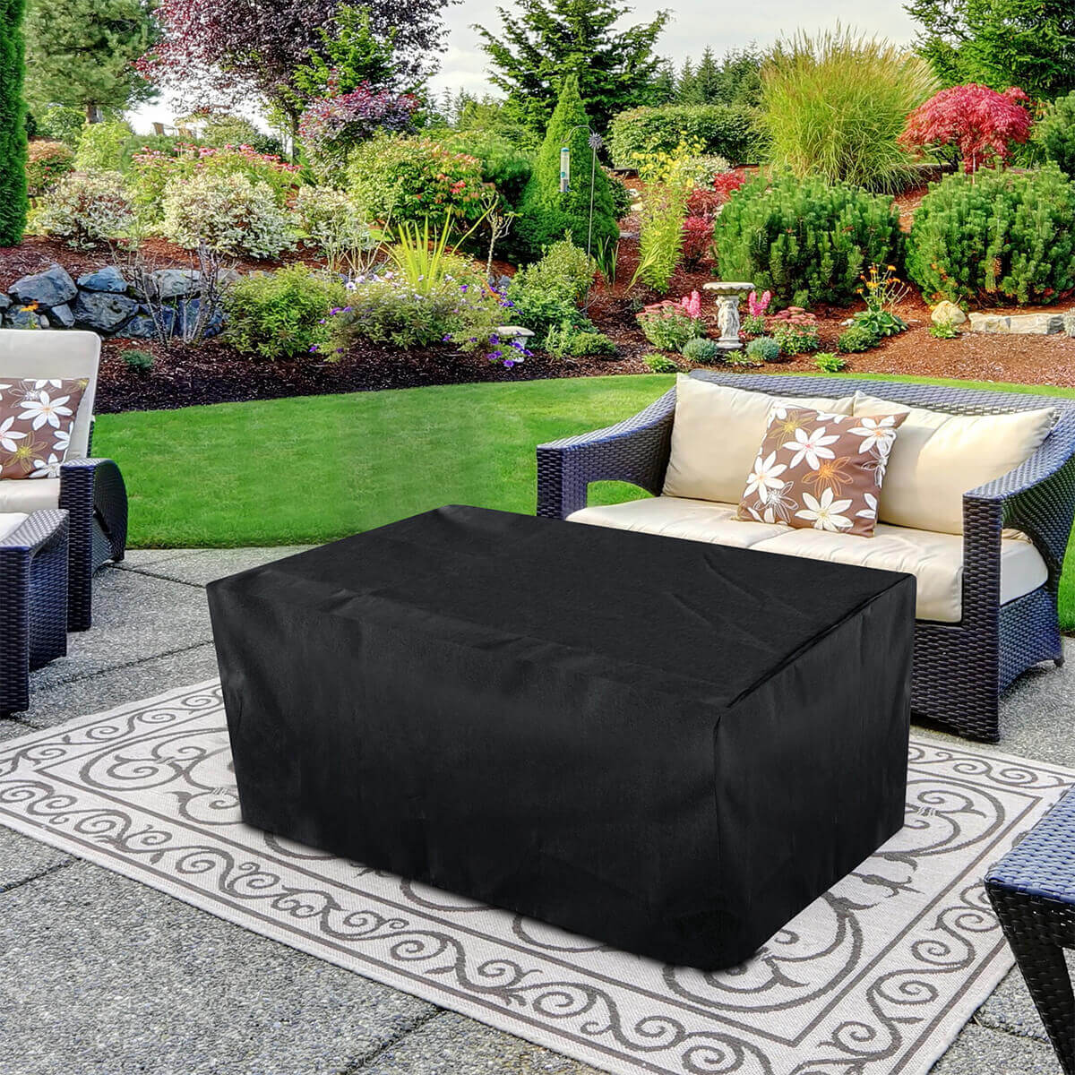 A Sunbeam Serenity Fire Table fully covered with the included black protective cover. The cover drapes neatly over the rectangular form, hiding all table surfaces including the slatted tabletop, burner area, and base. The fabric has a smooth matte texture with soft folds and light creases indicating a flexible but durable weather-resistant material. The cover reaches the ground on all sides and its shape clearly outlines the table’s wide top and straight-sided base. The covered table stands on a patterned outdoor rug between wicker seating, with a lush landscaped garden featuring trimmed shrubs, varied foliage, and layered greenery extending across the background.