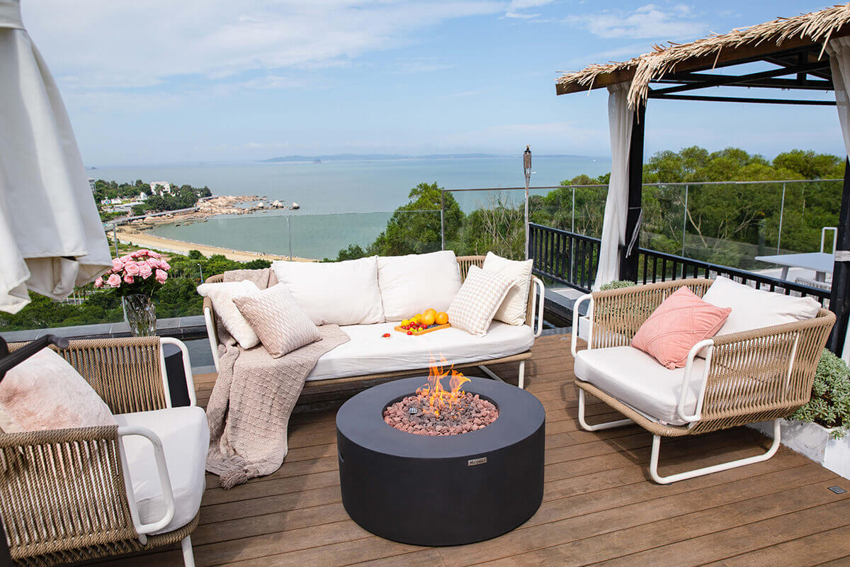 A rooftop lounge scene featuring the black Modeno Venice Fire Table centred between woven outdoor armchairs and a cushioned three seat outdoor sofa. The round fire table has a smooth glass fibre reinforced concrete body in matte black with a wide flat rim and an open circular burner filled with reddish brown and charcoal grey lava rock. A steady flame rises from the centre of the lava rock. The setup overlooks a coastal shoreline with a sandy beach, green tree canopy and calm ocean water beyond a glass railing. Cushions, throws, a vase of pink roses and a tray of assorted fruit decorate the outdoor seating area.