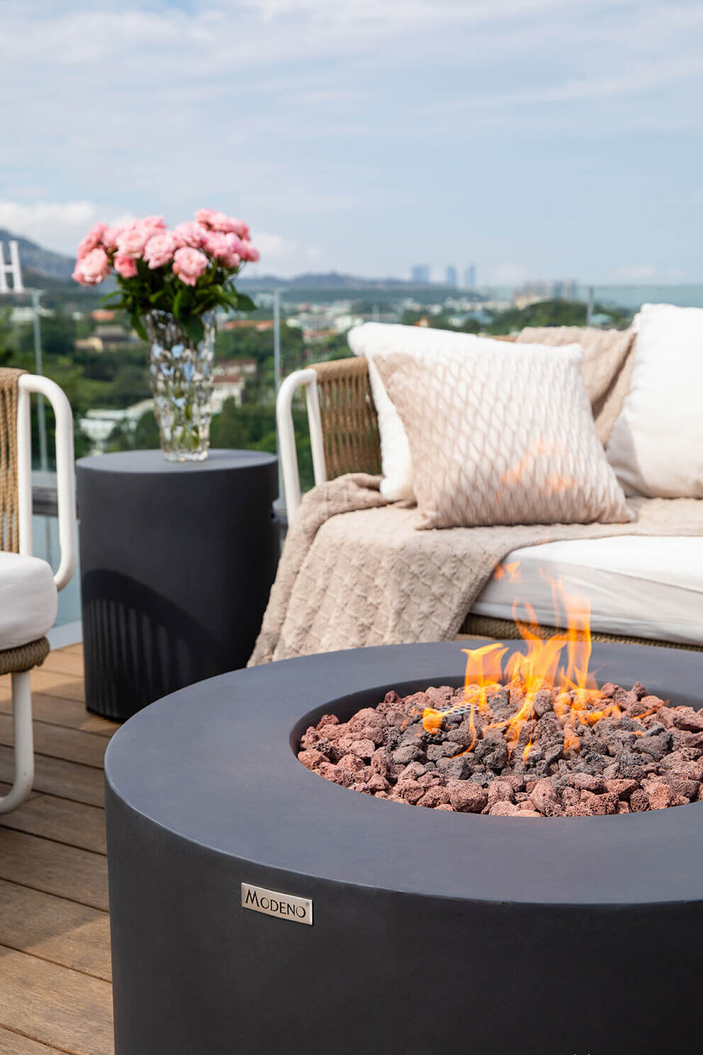 A close view of the Venice Fire Table in a rooftop setting, focusing on the upper rim and central burner filled with coarse lava rock in red and deep grey tones. The flame dances upward from the centre while the polished metal Modeno badge is visible on the front of the fire table’s smooth black concrete surface. In the background, soft throw pillows, a textured blanket and a tall vase of pink roses sit atop an outdoor side table. The distant cityscape and coastline appear slightly blurred beyond the glass railing.