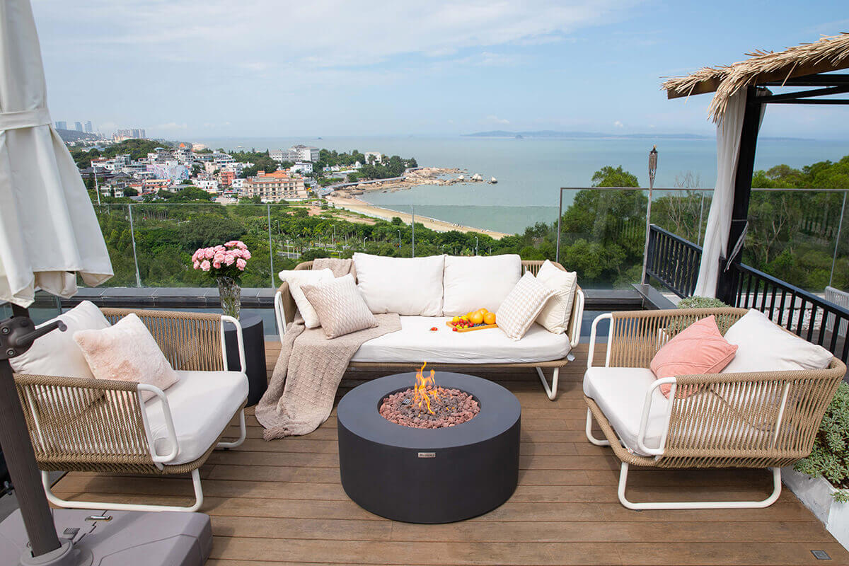 A wide rooftop patio arrangement with the Venice Fire Table placed at the centre of an outdoor seating area consisting of a white cushioned sofa and two woven armchairs. The round black concrete fire table holds a bed of mixed lava rock with flames rising from the burner. A tray with oranges and cherry tomatoes rests on the sofa. The background shows the shoreline, low rise buildings, lush greenery and calm ocean water under a hazy sky.