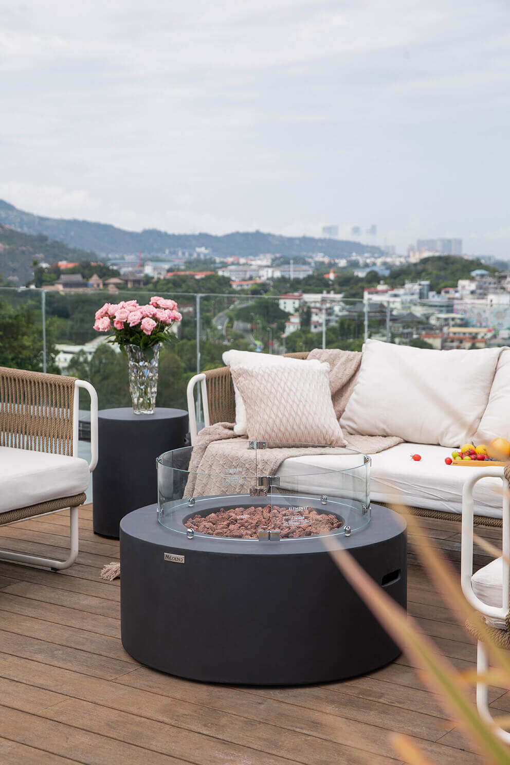 The Venice Fire Table is shown with a circular glass windscreen installed around the burner. The windscreen is made of curved glass panels held by polished metal brackets, encircling the lava rock bed inside the wide concrete rim. Small orange flames rise inside the protected burner area. Surrounding the fire table is an outdoor seating area with woven chairs, white cushions, a textured knitted throw, fruit on the sofa and a tall vase filled with pink roses. Beyond the glass railing, a distant view of city buildings, rolling landscape and cloudy sky can be seen.