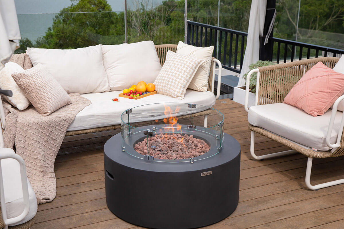 A closer side view of the Venice Fire Table with the circular glass windscreen attached around the burner. The lava rock bed fills the recessed opening inside the table’s thick black concrete rim. Soft orange flames are visible behind the windscreen. To the left and right, cushioned outdoor chairs frame the setting, and a glass vase of pink roses stands on a matching side table. In the background, the coastline, green treetops and distant rooftops stretch toward the horizon.
