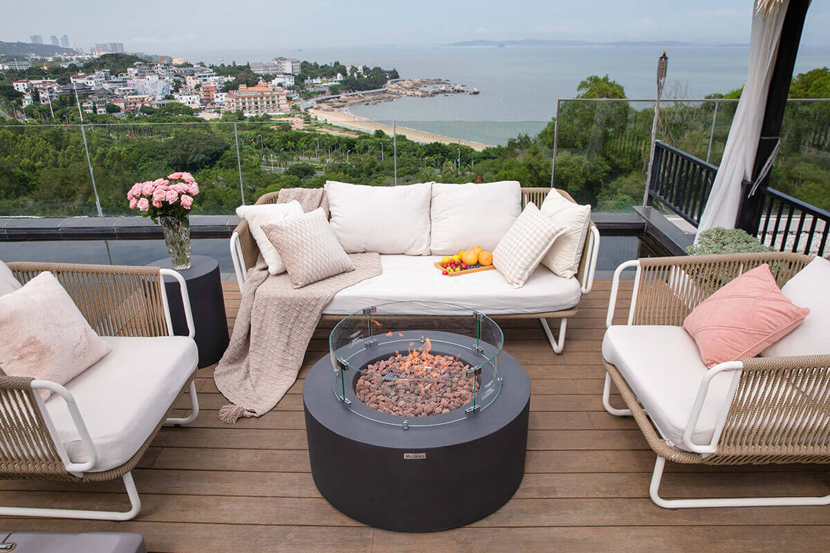 Another rooftop patio scene showing the Venice Fire Table with the glass windscreen surrounding the burning lava rock. The flames are contained neatly within the circular windscreen. The seating area includes a cushioned sofa with textured pillows and a knitted throw, flanked by woven chairs with white cushions. A glass vase of pink roses sits to the left, while a tray of oranges and cherry tomatoes rests on the sofa. Through the clear patio railing, the coastline, beach, treetops, and ocean extend into the distance.